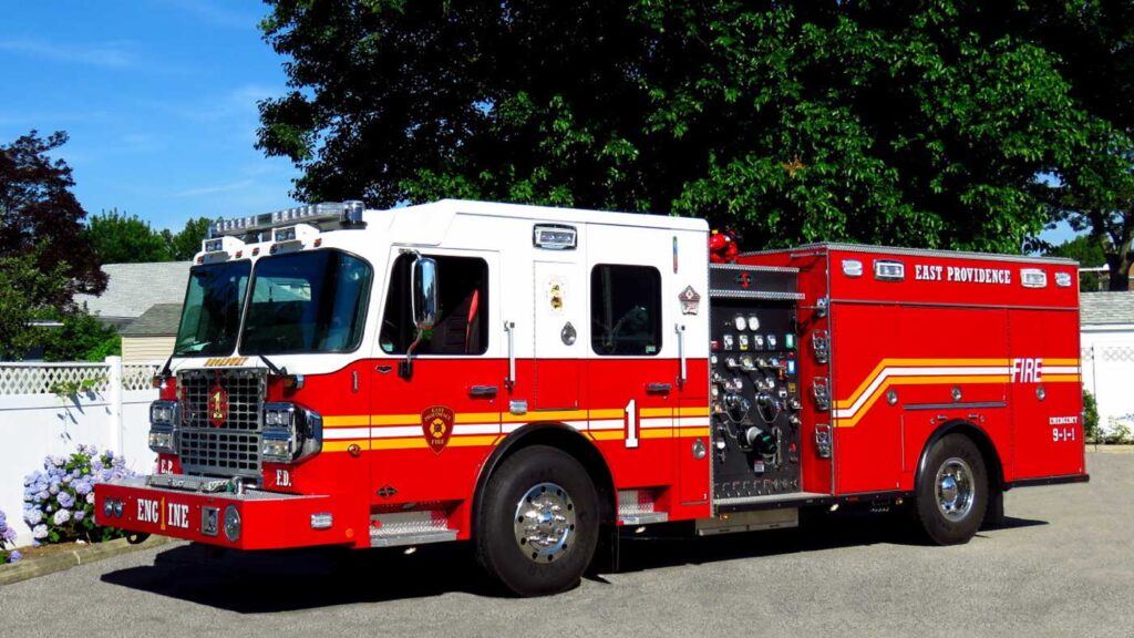 A red and white fire truck marked "East Providence" is parked on a paved area with trees and houses in the background under a clear sky.