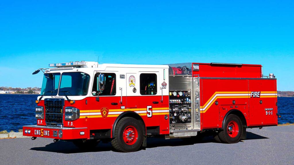 A red and white fire truck with the number 5 on the side is parked on a paved surface near a body of water under a clear blue sky.