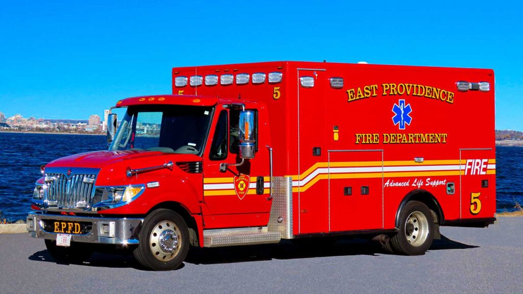 A bright red East Providence Fire Department ambulance is parked by a waterfront on a sunny day, with city buildings visible in the distant background.