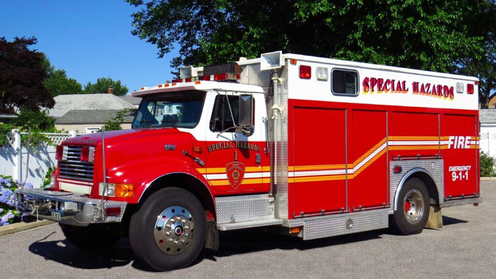 A red and white “Special Hazards” fire rescue truck is parked on a sunny street near houses and trees.