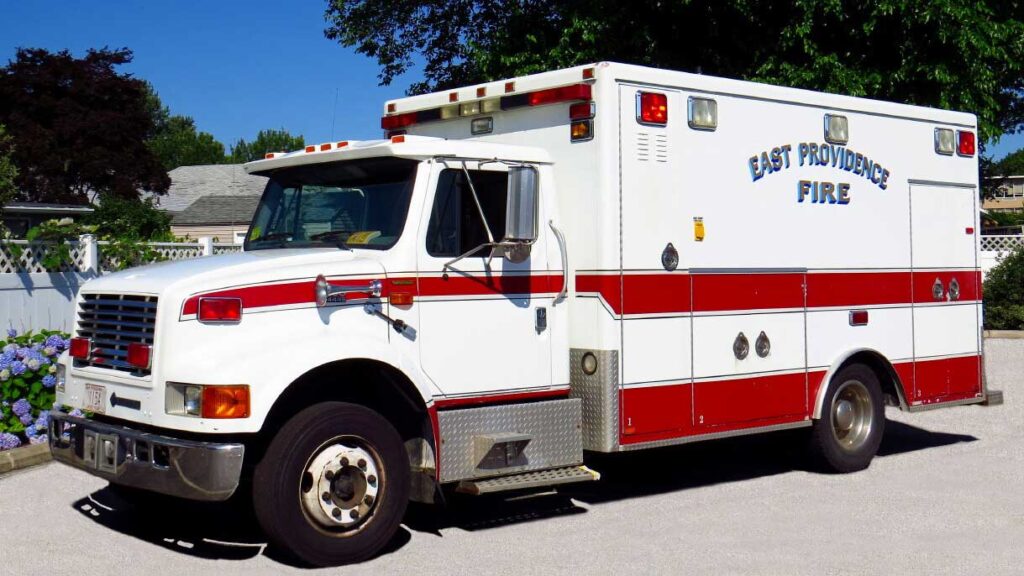 A white and red ambulance with "East Providence Fire" written on the side is parked on a sunny street near a fence and flowers.