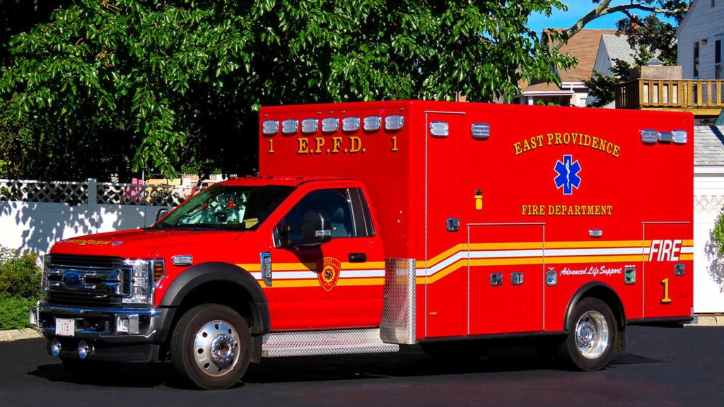 A red East Providence Fire Department ambulance is parked on pavement near houses and trees on a sunny day.