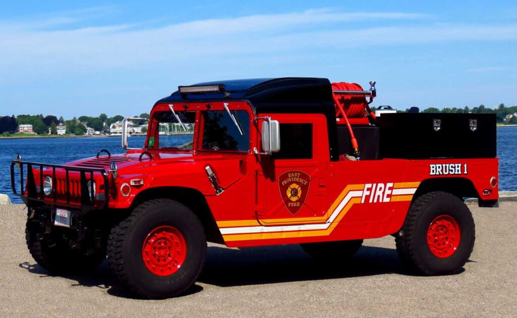 A red fire department brush truck is parked near water on a sunny day. The vehicle has large tires, firefighting equipment, and “FIRE” with yellow stripes on the side. Houses and trees are visible in the background.