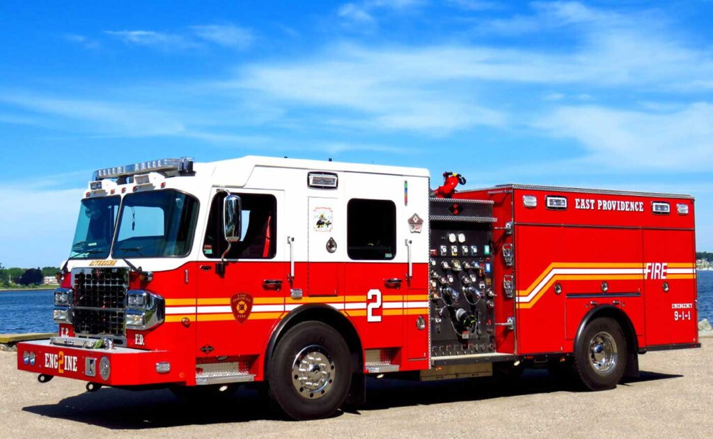 A red and white fire truck labeled “East Providence Fire” is parked on pavement near water under a blue sky with light clouds.