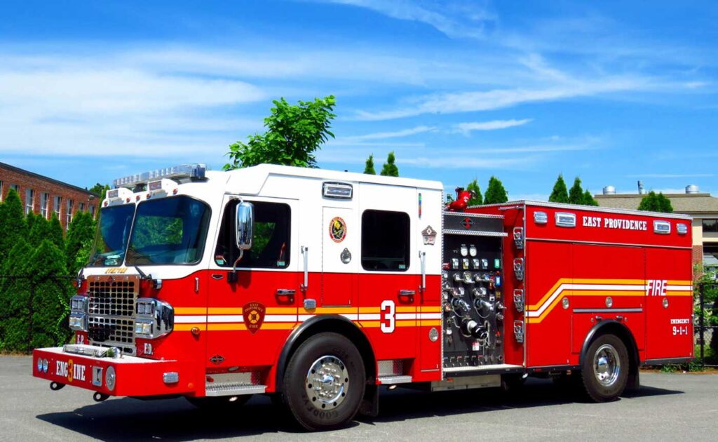 A bright red fire truck labeled “East Providence” and “Engine 3” is parked outdoors on a sunny day, with trees and buildings visible in the background.