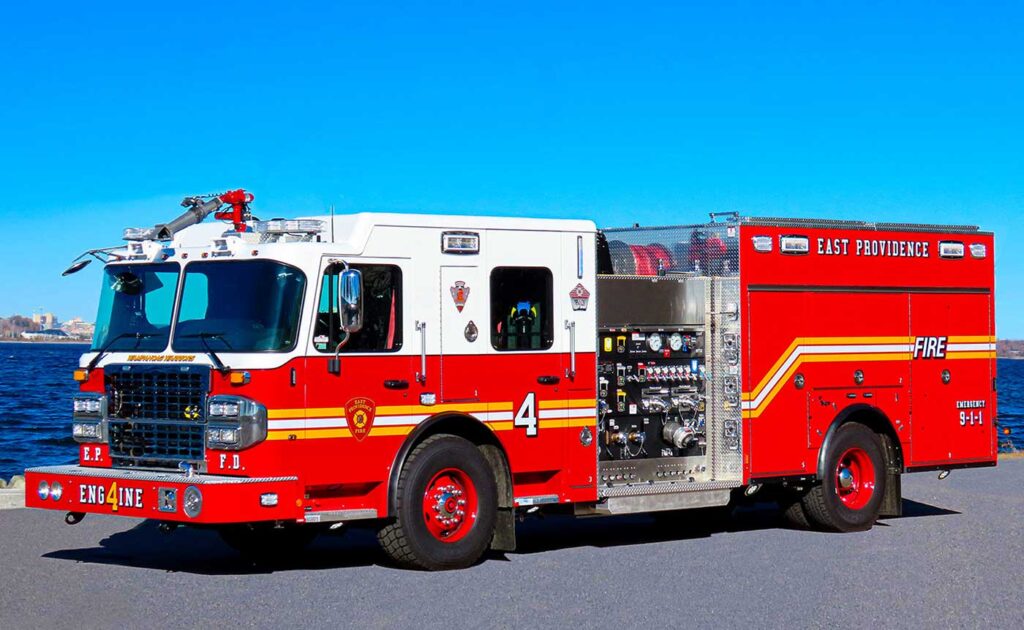 A red and white fire truck labeled “Engine 4” and “East Providence” is parked on a paved area near water, with a clear blue sky in the background.