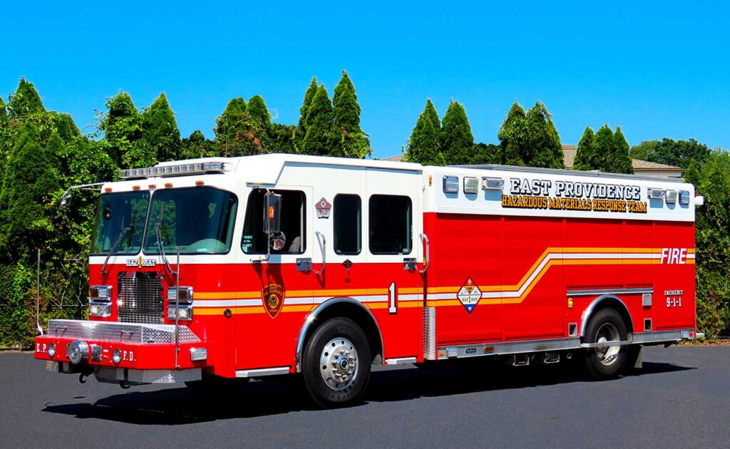 A red and white fire truck labeled “East Providence Hazardous Materials Response Team” is parked on a blacktop road with green trees in the background.