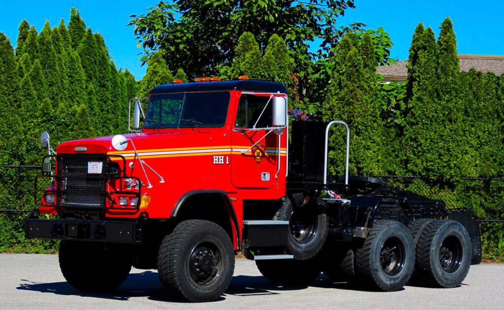 A bright red fire department heavy rescue truck with six large wheels is parked on pavement, surrounded by green trees and a fence under a clear blue sky.