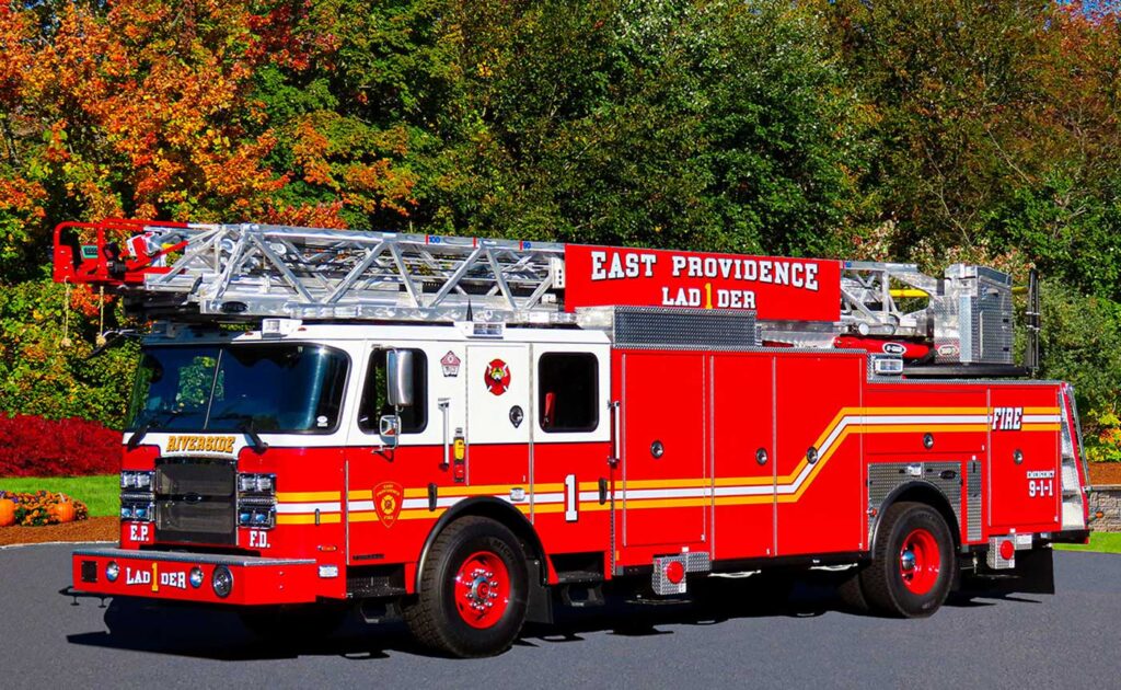 A bright red fire truck with an extended ladder, marked “East Providence Ladder,” is parked on a paved road with trees and bushes in the background.