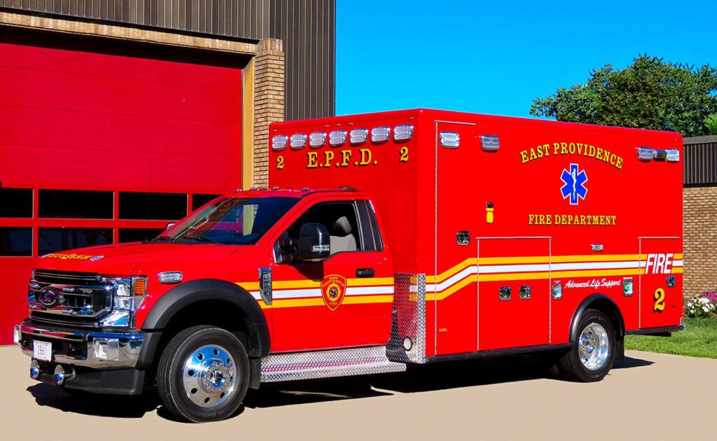 A red East Providence Fire Department ambulance is parked outside a building with red garage doors. The vehicle has yellow and white stripes and emergency medical symbols on the side.