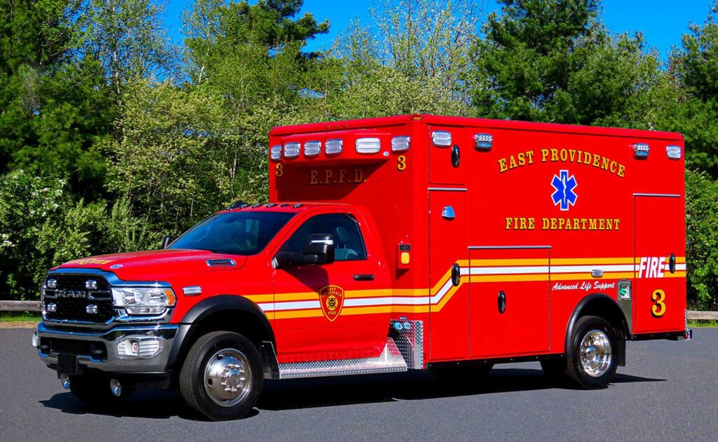 A bright red East Providence Fire Department ambulance is parked on a road, with trees and greenery in the background. The vehicle features yellow stripes and emergency symbols.