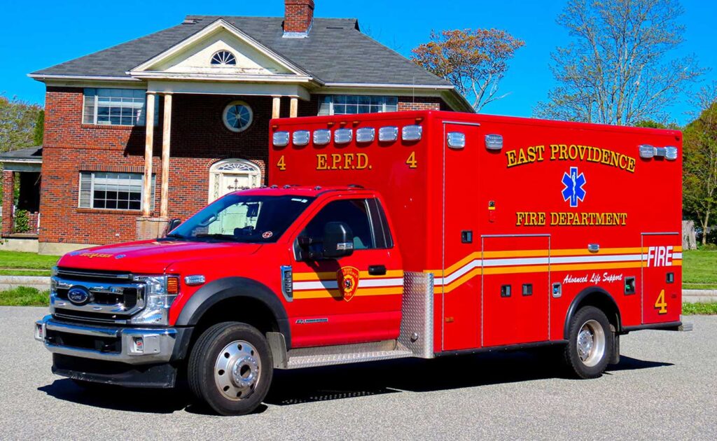A bright red East Providence Fire Department ambulance is parked on a street in front of a large brick house with white columns and a black roof, on a sunny day with a clear blue sky.
