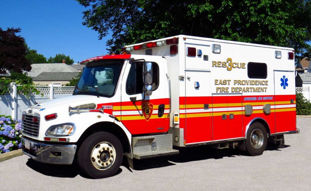 A red and white East Providence Fire Department rescue ambulance is parked on a sunny street, with trees and houses in the background.