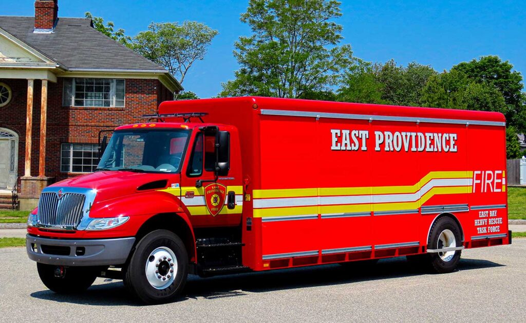 Bright red East Providence fire rescue truck parked on a street, with yellow and white stripes, “East Providence” and “FIRE” written on the side. There are trees and a brick house in the background.