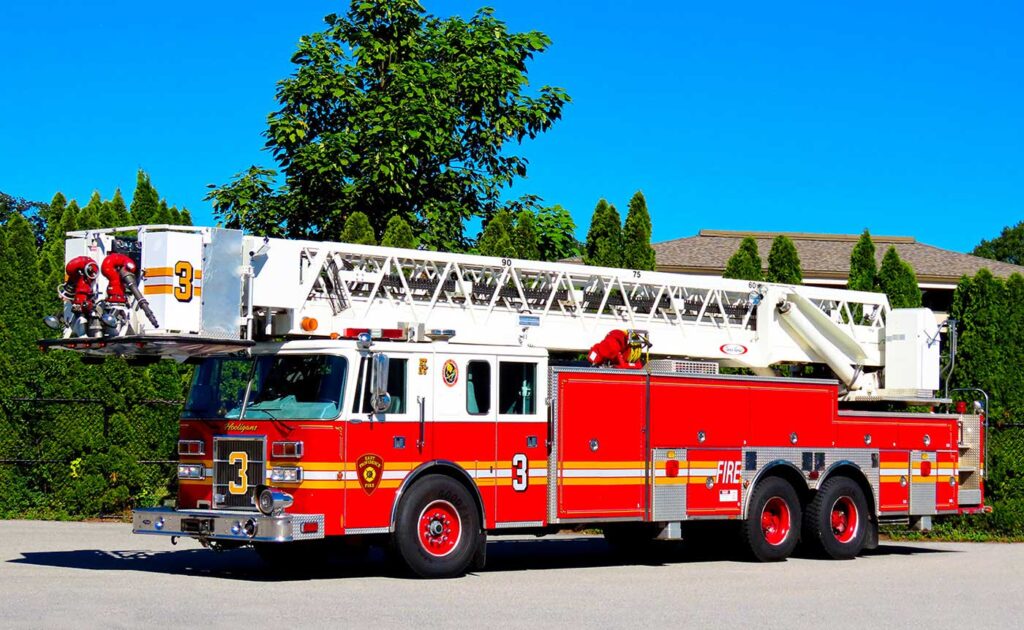 A red and white fire truck with an extended ladder is parked on a sunny street, with green trees and bushes in the background.