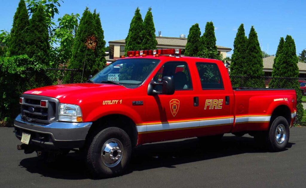 A red fire department utility truck is parked on pavement in front of tall green trees. The truck has emergency lights on top and “FIRE” written on the side.