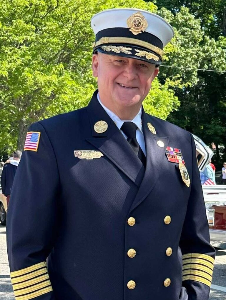 A smiling fire chief in full dress uniform stands outdoors near parked cars and people, with trees in the background.